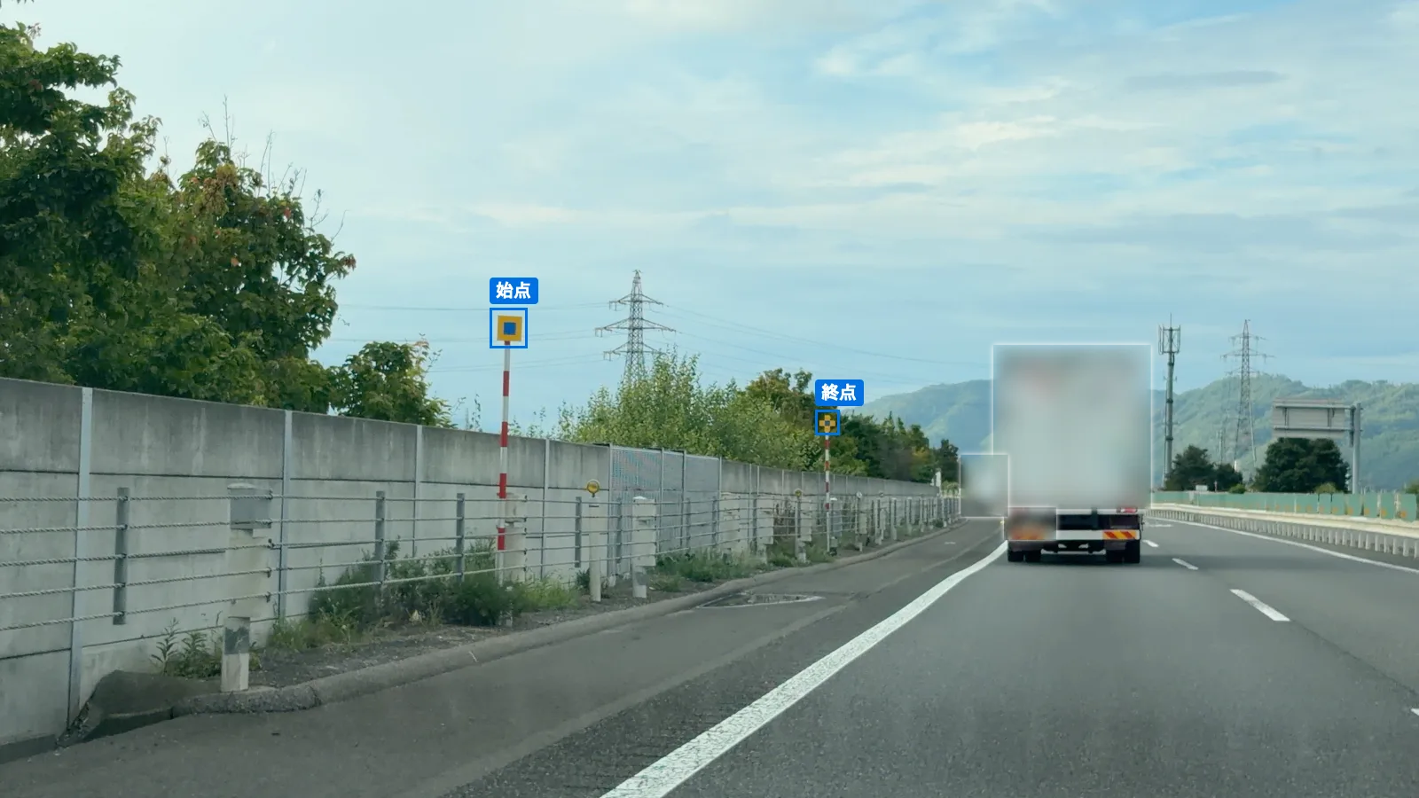 高速道路の左の路肩に立つ投雪防止表示板。手前の始点(黄色の正方形の中に青い正方形)と、遠くの終点(黄色と青の十字形)が青い枠で囲まれている。前を走るトラックはぼかしてある